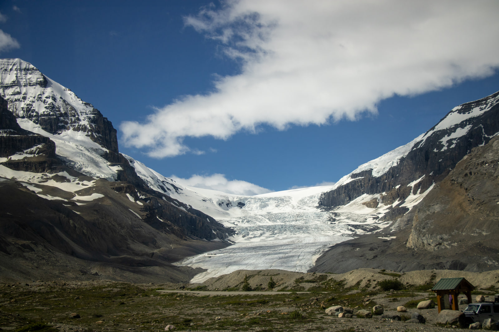 A glacier tongue comes down from a broken icefall between a snowy peak and a snow-topped cliff, and a green-roofed kiosk and grey SUV stand at lower right.