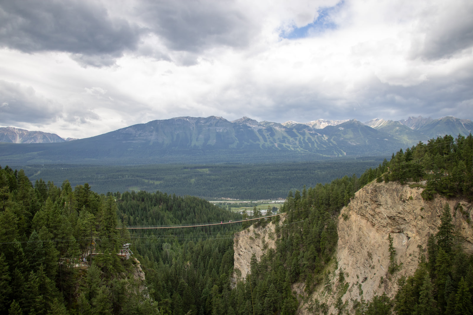 Tiny people cross a thin walkway above dense forest and a pale cliff, and a mountain marked with ski runs sits across a wide valley under heavy cloud.