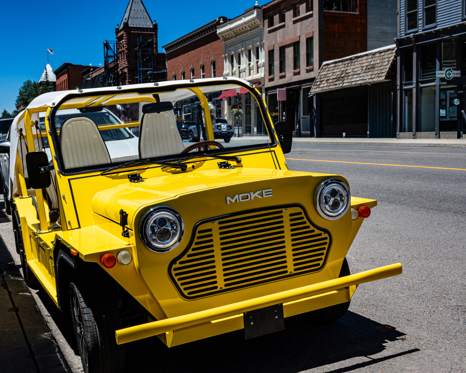 An open-sided yellow car with a white canvas roof is parked across from brick storefronts, with a sandstone tower in scaffolding down the street.