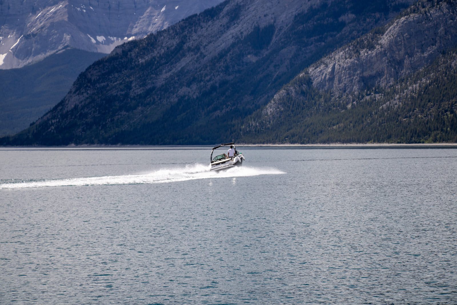 A white motorboat trails a long line of spray on rippled grey water, below a steep forested mountainside in shade and a grey peak with snow patches.