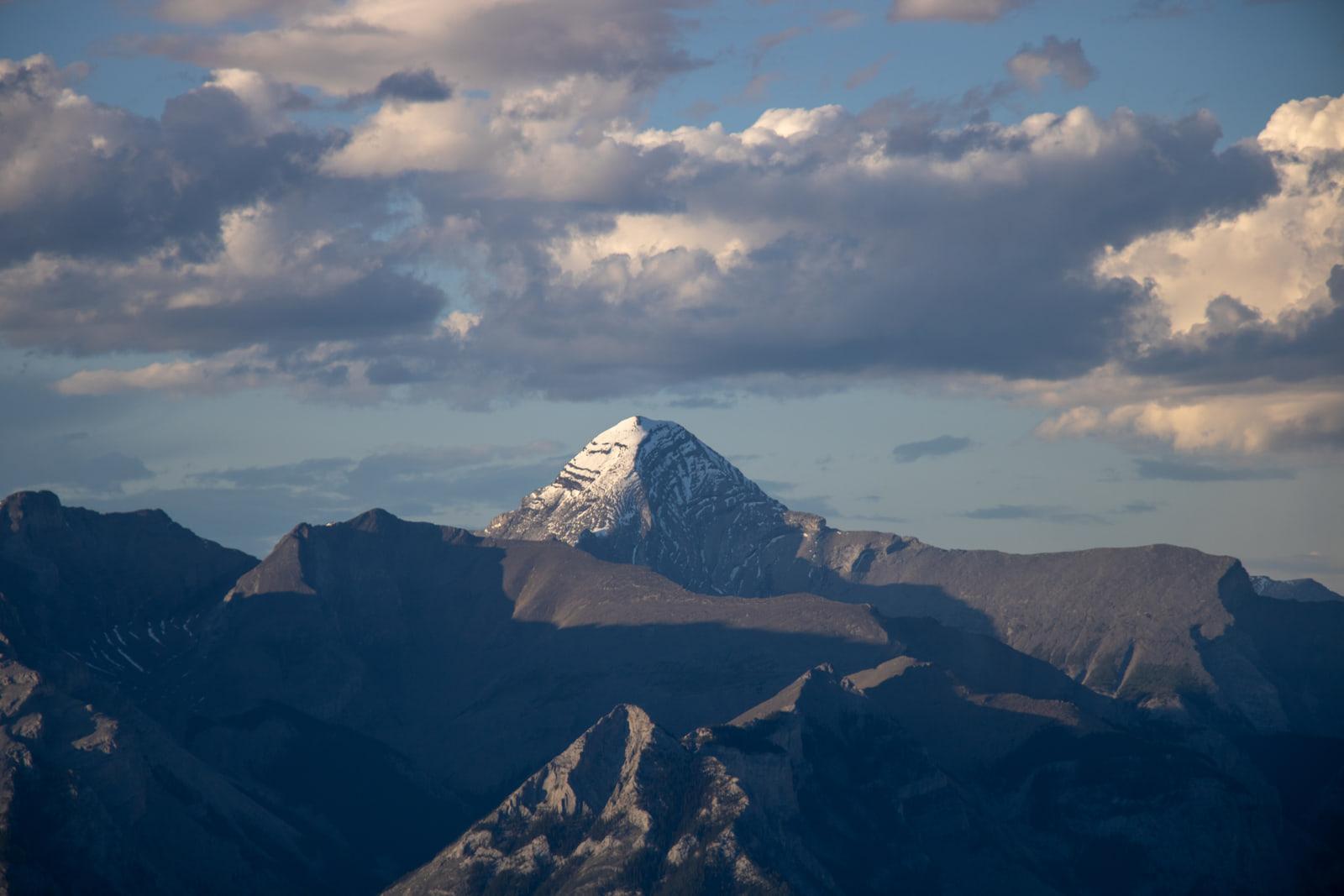 A sunlit snow-capped pyramid peak stands against dark blue storm clouds, with two shadowed mountain ridges layered in front of it.