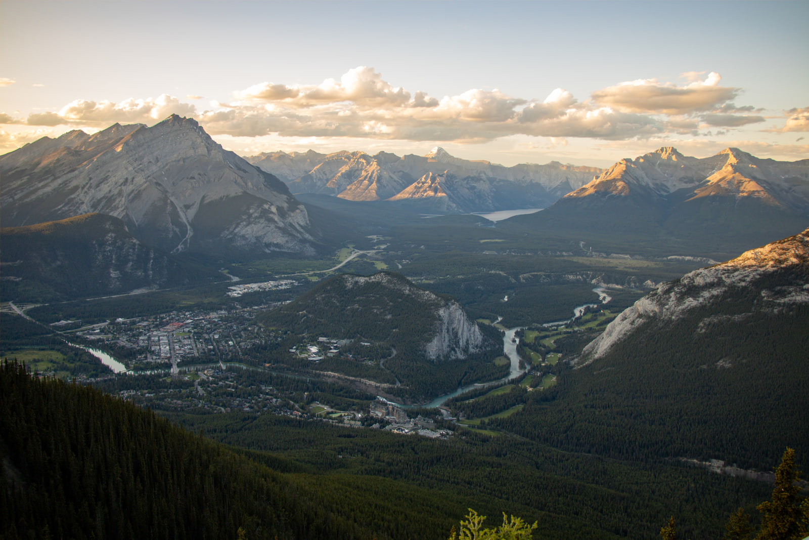 A town and a curving river lie on a dark, forested valley floor below a big grey mountain, with warm light on the far peaks and clouds.