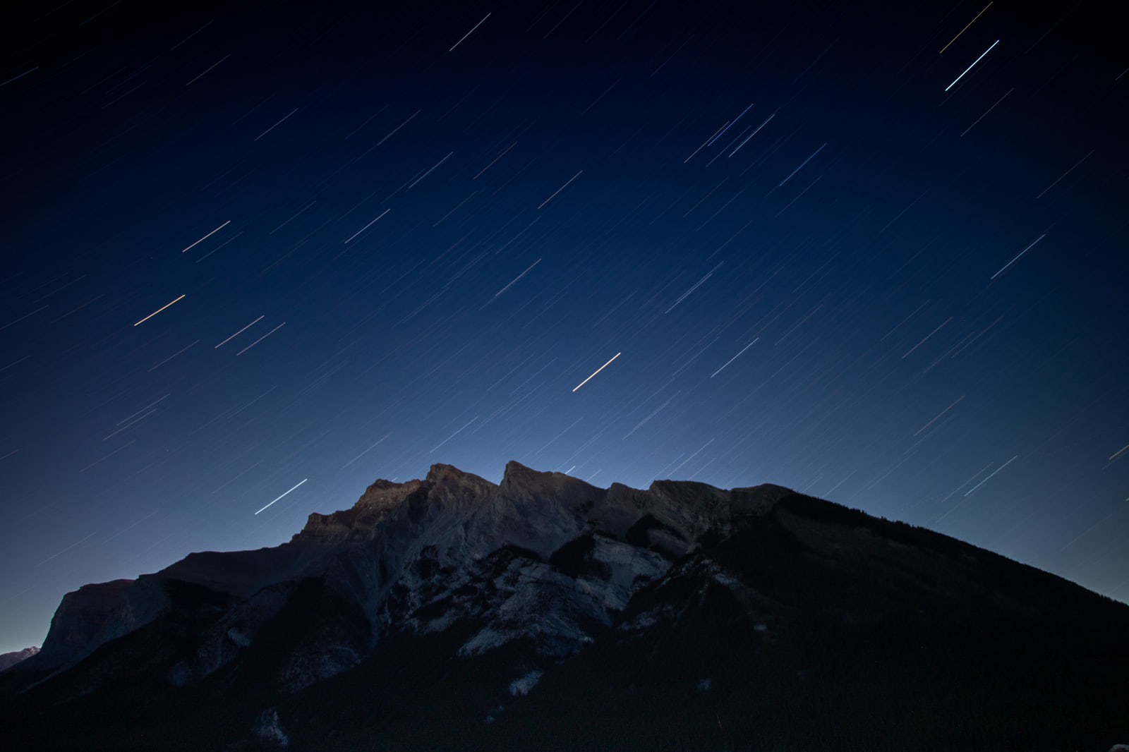 A mountain with a jagged ridge, faintly lit grey slabs and dark forested lower slopes sits under a navy sky crossed by short diagonal star streaks.